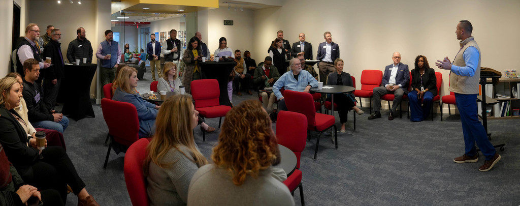 A group of people in a room with chairs and tables. One person stands and speaks to the audience.