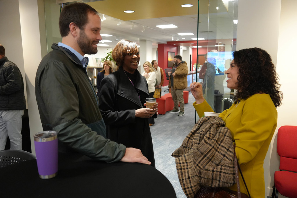 A man and two women in coats converse in an office space with a red chair and table.