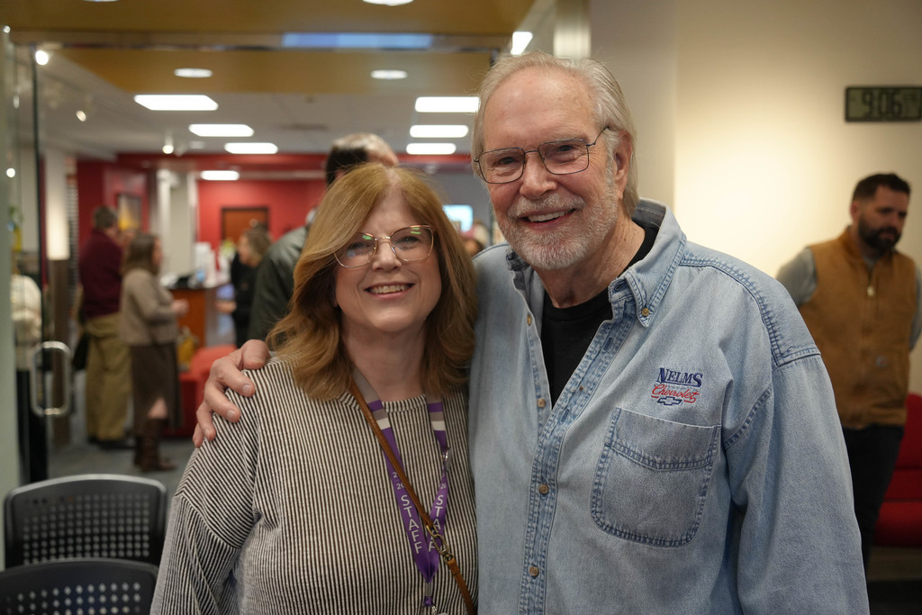 A man and woman stand close, smiling, in a hallway. Behind them, people walk and sit in chairs.