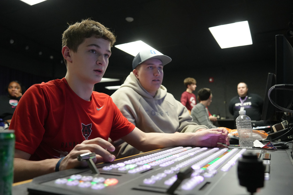Two young men seated, one wearing a red shirt, the other a gray hoodie and cap. They focus on a console with buttons.