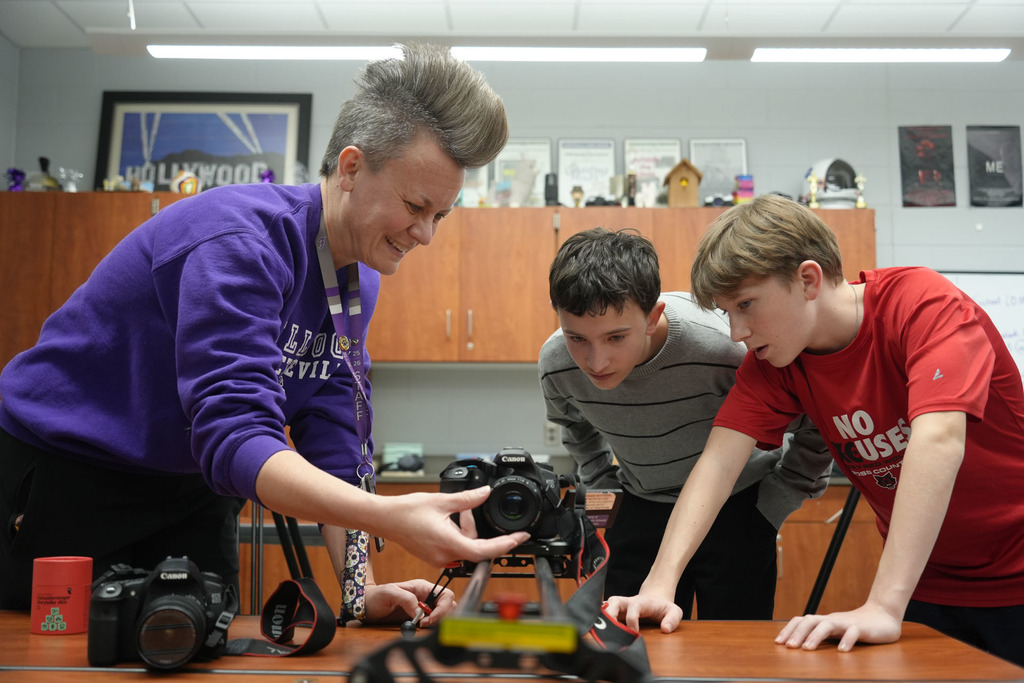 A woman and two boys lean over a table examining equipment. Behind them, shelves display various items.