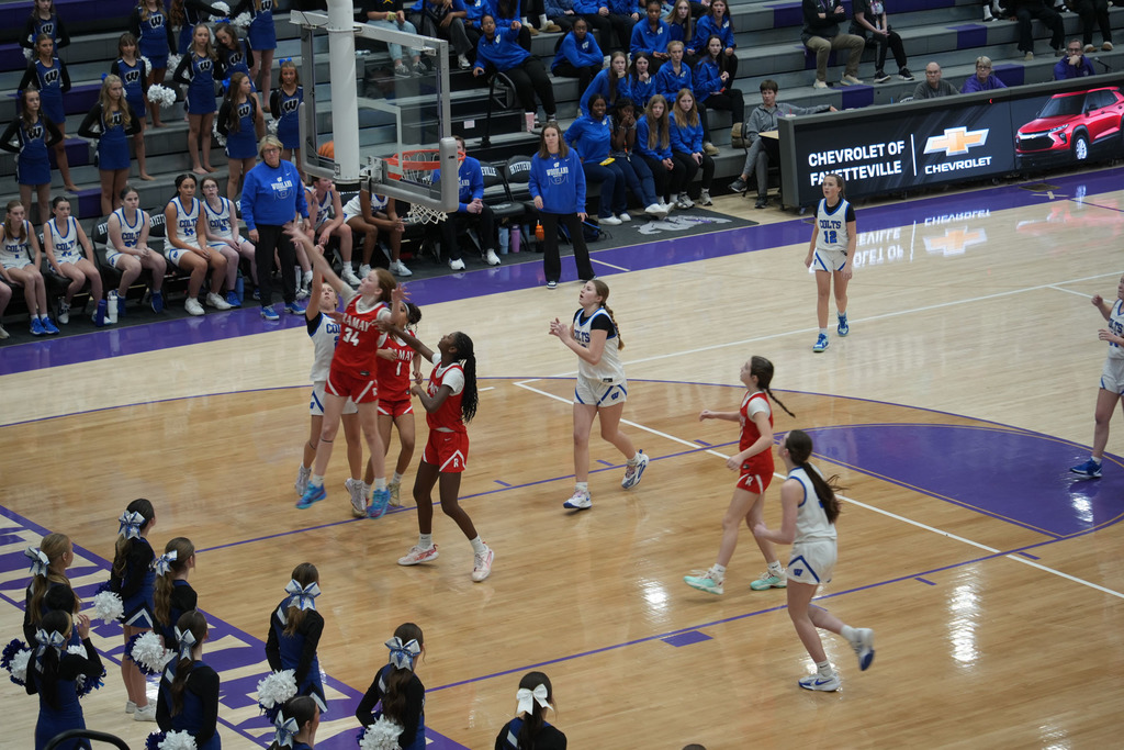 Basketball players on a court, some celebrating, others running. Spectators in blue sit on bleachers. A red car ad is on the wall.