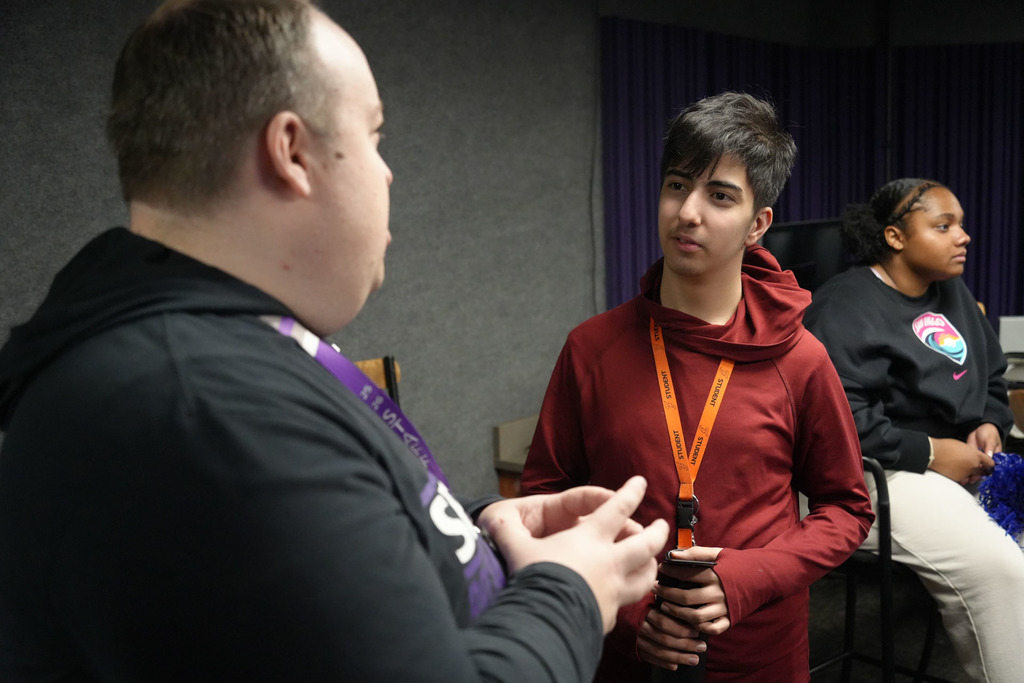 Two individuals face each other; one in a maroon hoodie speaks while the other listens. A woman sits behind them with a colorful lanyard.