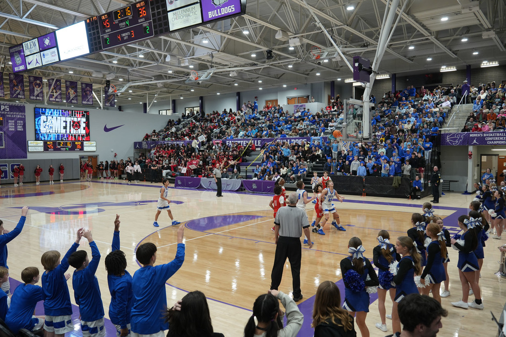 An indoor basketball game with players, a referee, and cheerleaders in blue uniforms. Spectators are seated on bleachers.