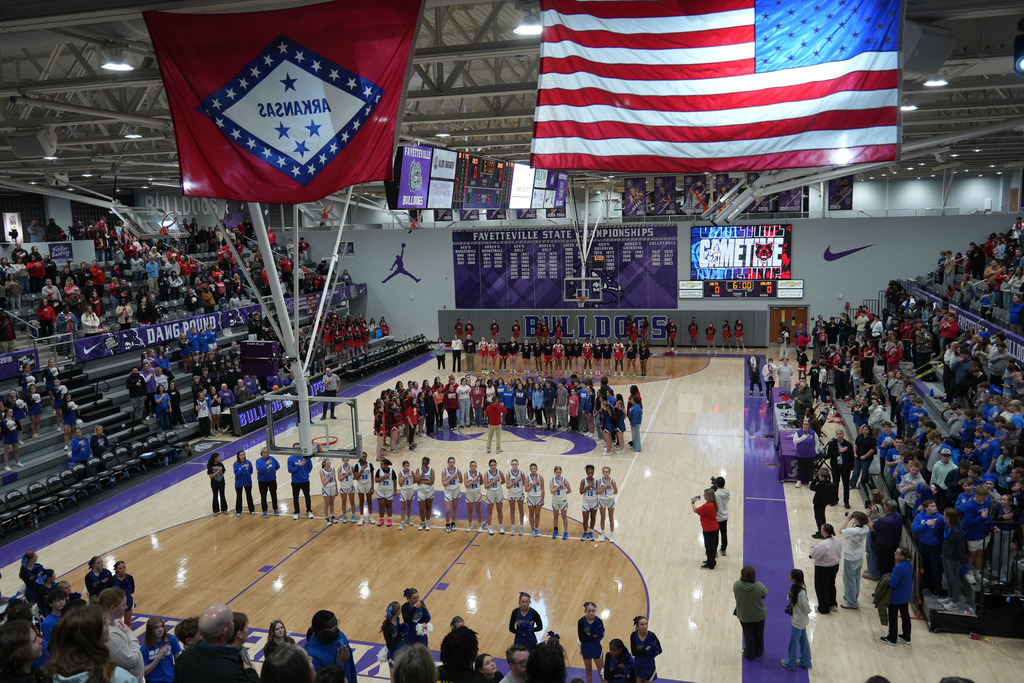 Basketball court with a crowd of people. Two flags hang from the ceiling, one featuring a star design.