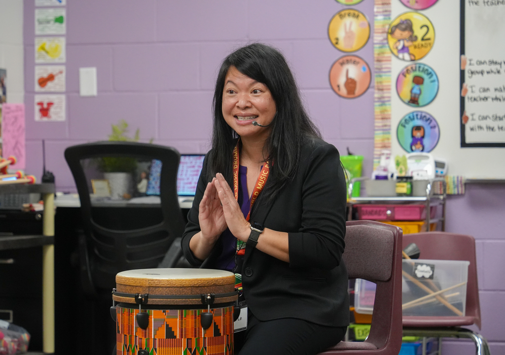 A woman claps near a drum in a classroom with purple walls and circular decorations.