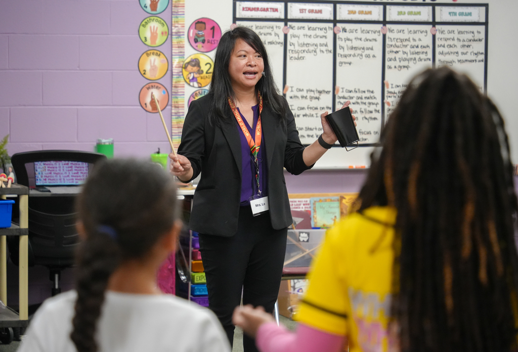 A woman speaks to two children in a classroom. A whiteboard with colorful icons is behind her.