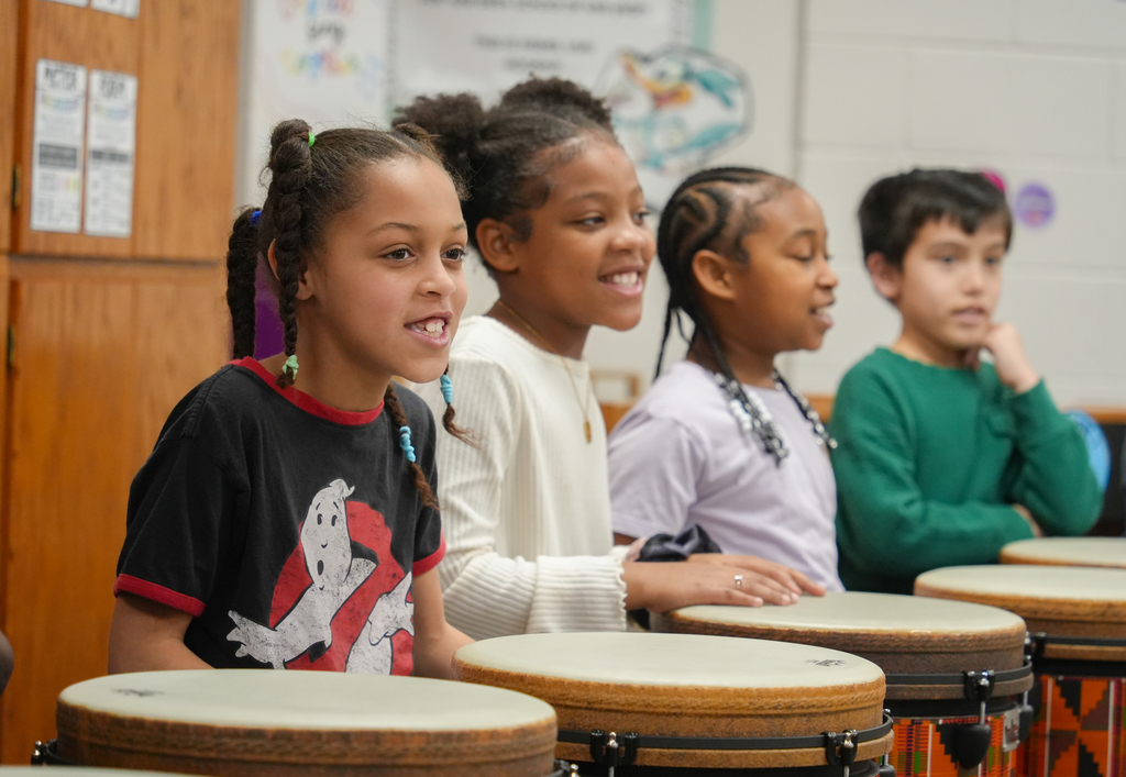 Four children seated in a classroom, playing drums. One smiles, another looks thoughtful, and two face the front.