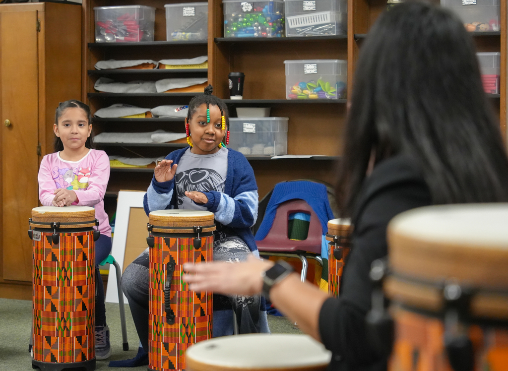 Three children seated in front of drums. A woman with long dark hair instructs them. Shelves filled with various items behind them.