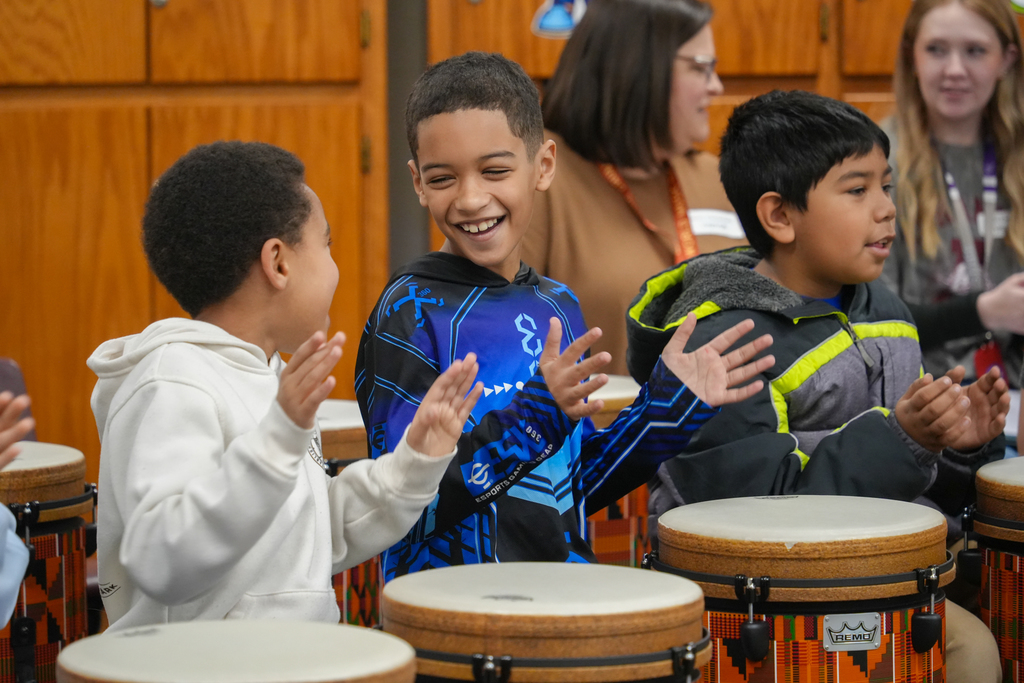 Three children clap and smile, with two playing drums in a classroom. A wooden cabinet and people stand in the background.