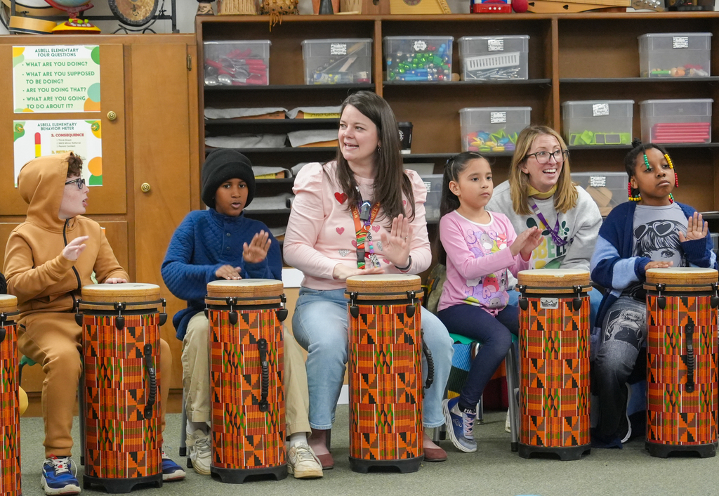 Five children and an adult woman seated around drums, smiling. The adult woman wears a pink shirt.