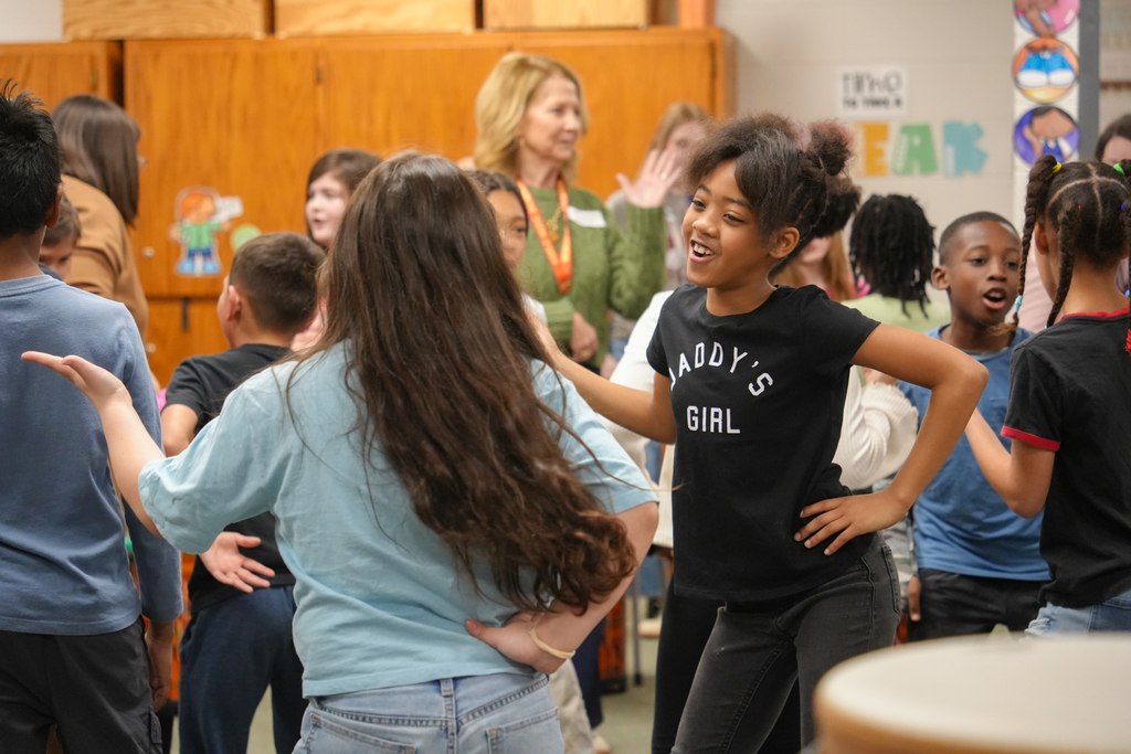 A group of children and adults in a classroom. A woman stands near wooden cabinets. Two girls dance, one in blue and one in black.