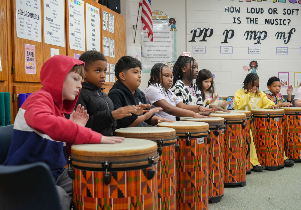 Children sitting in a classroom playing drums. Some are wearing hoodies. Wall has a flag and text.