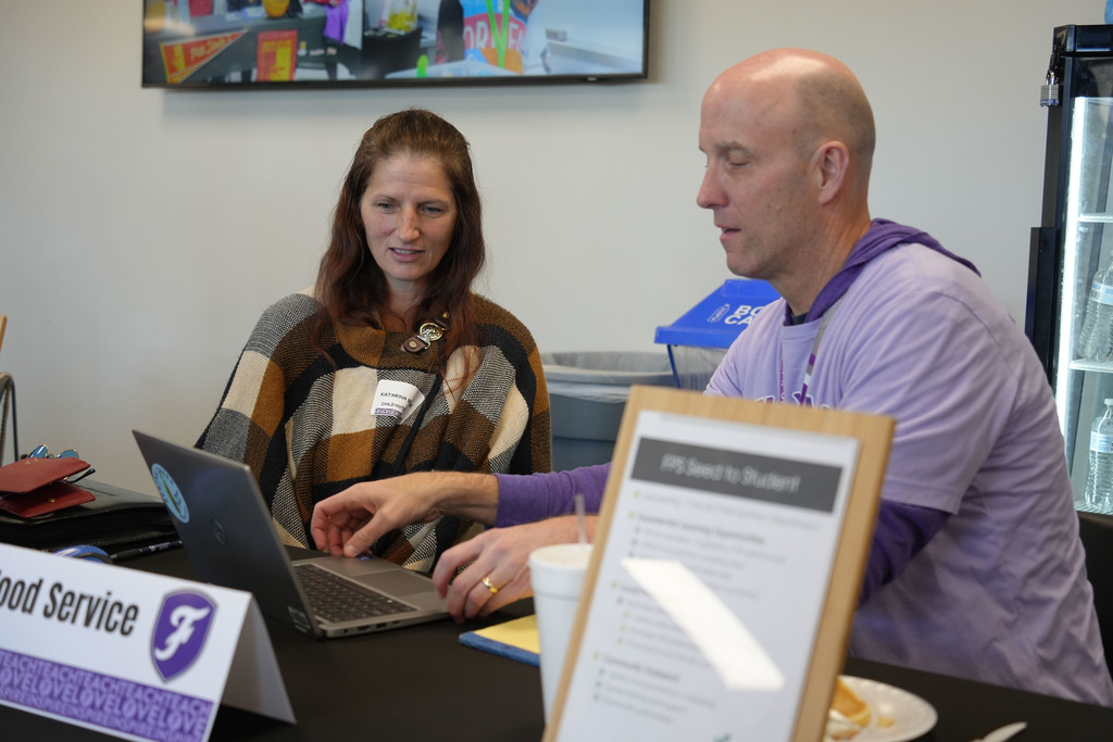 A man and a woman sit at a table, looking at a laptop. The man holds a paper.