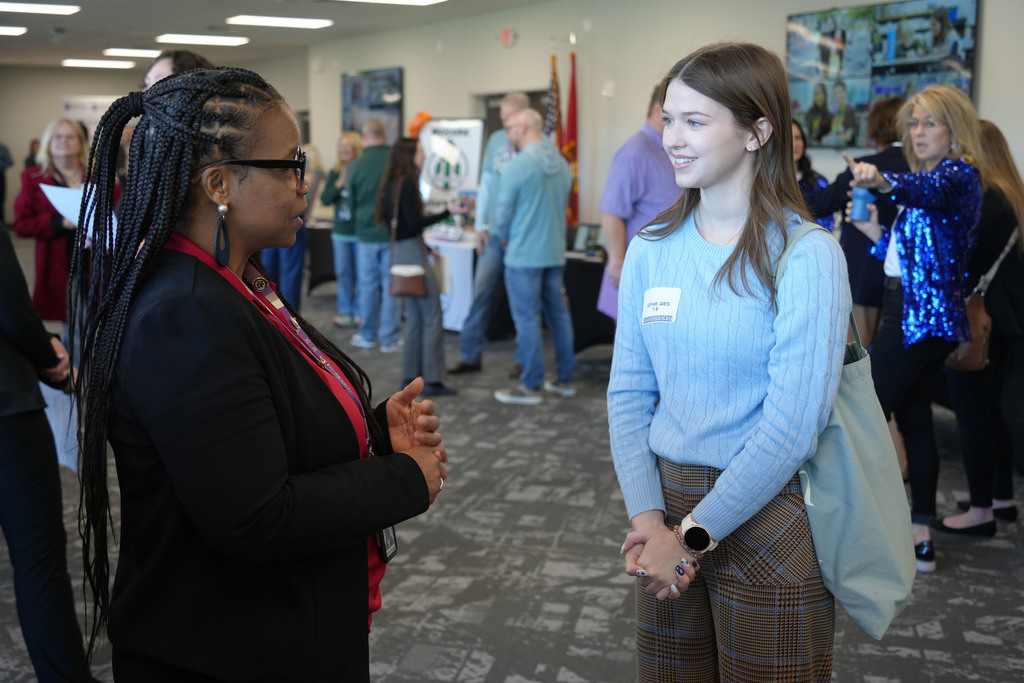 Two women talking at an indoor event, one in black, the other in blue, with a blurred background of other attendees.