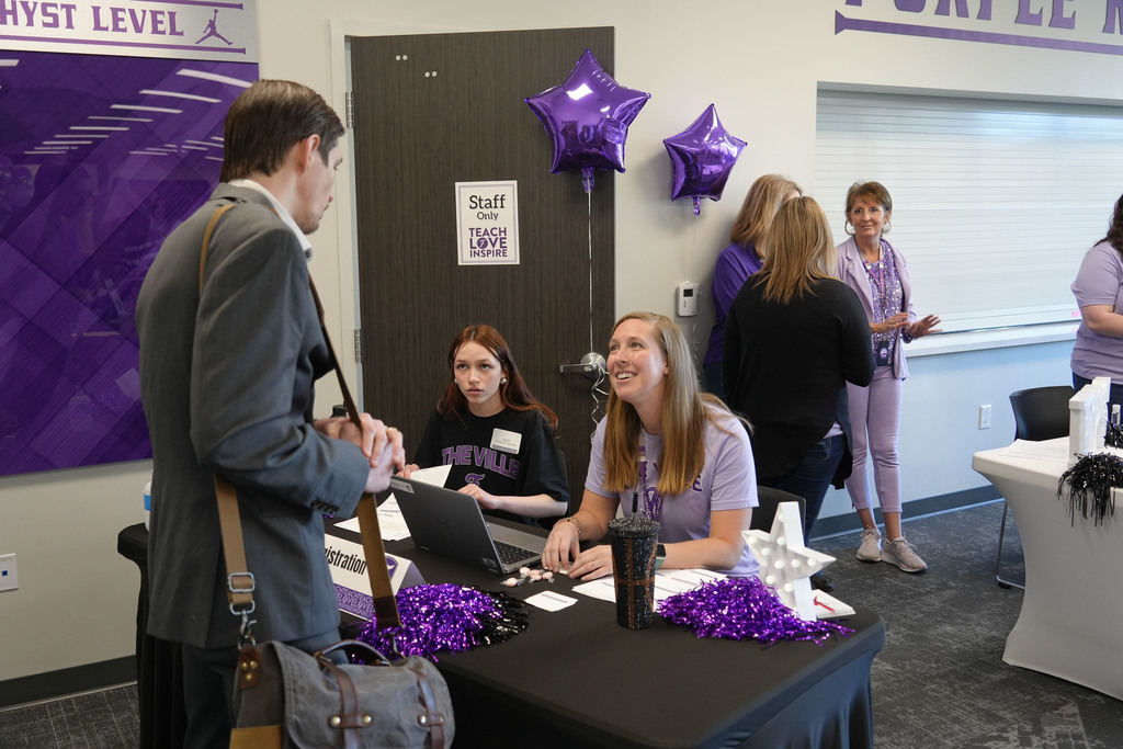 People at a table with purple balloons. A man talks to a woman at a laptop.
