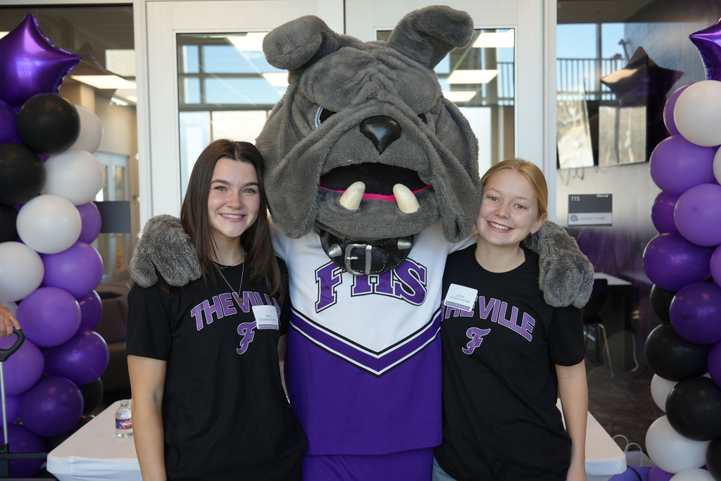 Two young women stand next to a mascot. The mascot wears a purple and white uniform. Behind them are balloons.