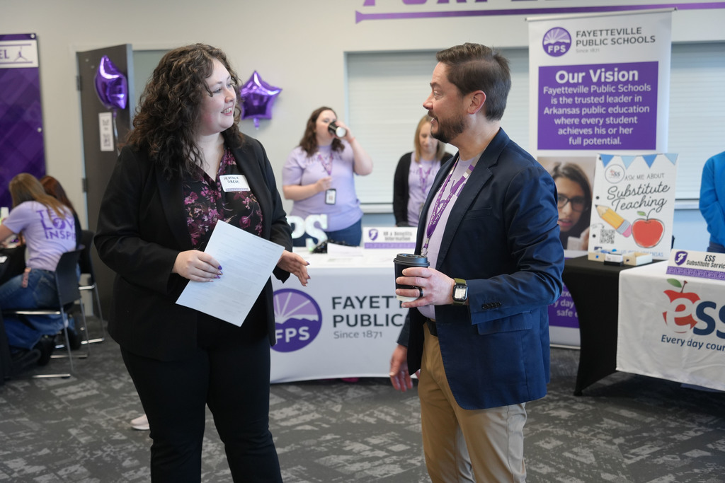 Two people in formal attire engage in conversation. A woman holds a piece of paper, and a man holds a cup.