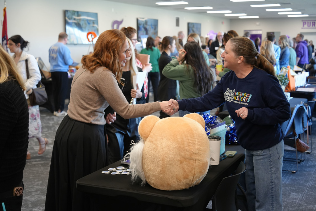 Two women shake hands next to a large stuffed bear on a table in a busy room.