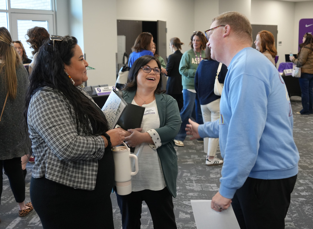 A group of people converses in a conference room. One woman holds a folder and mug, another holds a folder, and a man speaks to the woman.