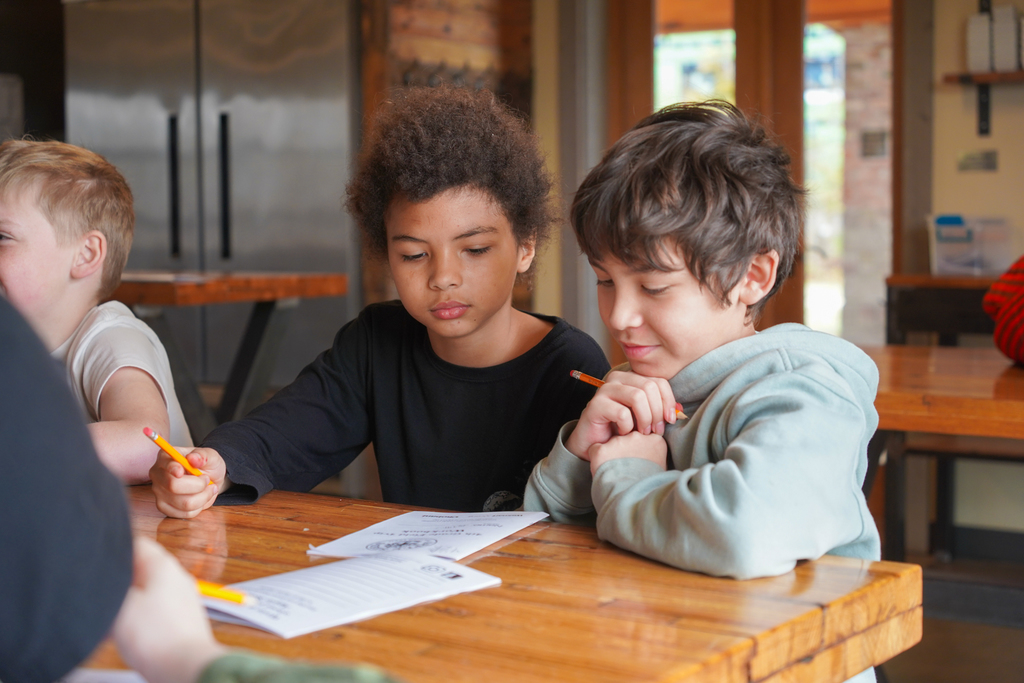 Two boys sitting at a table, one drawing with a pencil, the other looking at the drawing.
