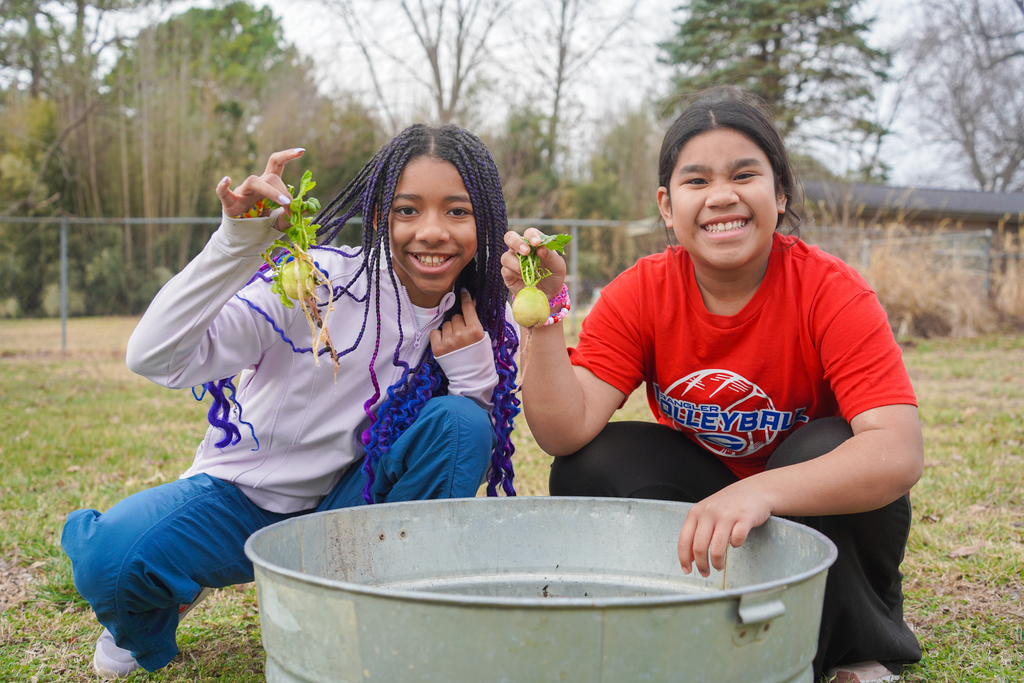 Two girls pose with vegetables, one holding purple beans. They sit beside a metal bucket on grassy ground, trees in background.