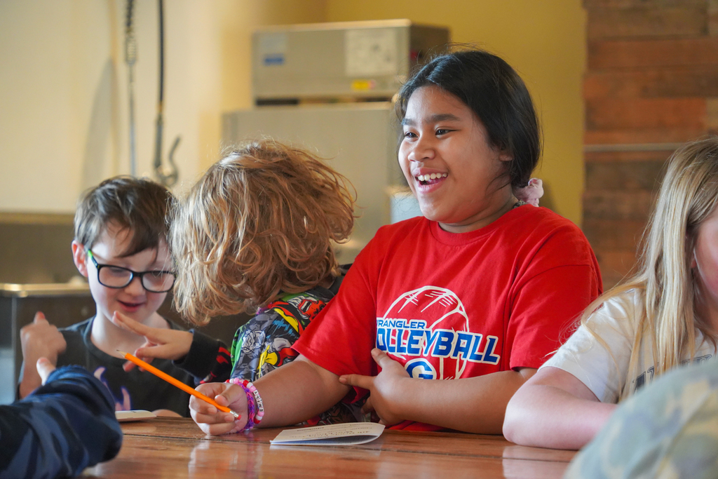 Kids in a classroom, one holding a pencil, another wearing glasses, and two smiling at each other.