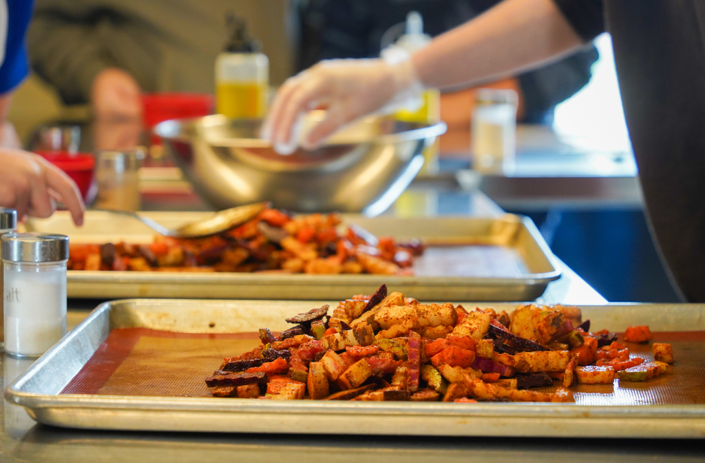 A kitchen with several trays of cooked food. A person in gloves mixes ingredients in a bowl.