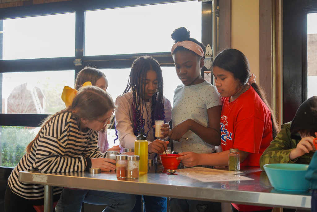 Group of children in a classroom around a table, preparing food, with bottles, bowls, and paper.