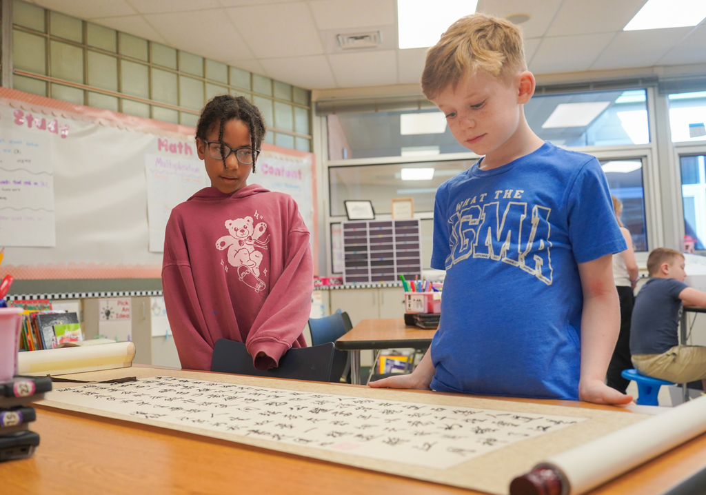 Two children, one in pink, the other in blue, study a scroll with Chinese characters on a desk in a classroom.