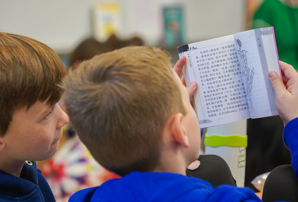 Two boys in a classroom, one holding an open book with foreign text, and the other looking at it.