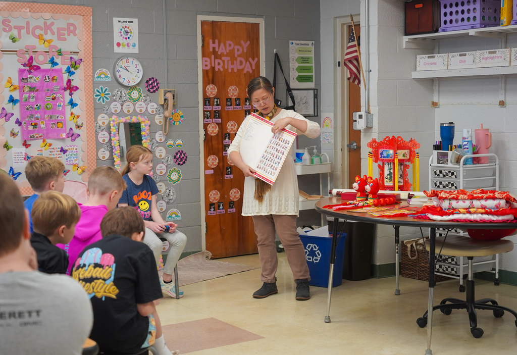 A teacher holds a large sheet of paper in a classroom, surrounded by children. Colorful decorations and posters adorn the walls.