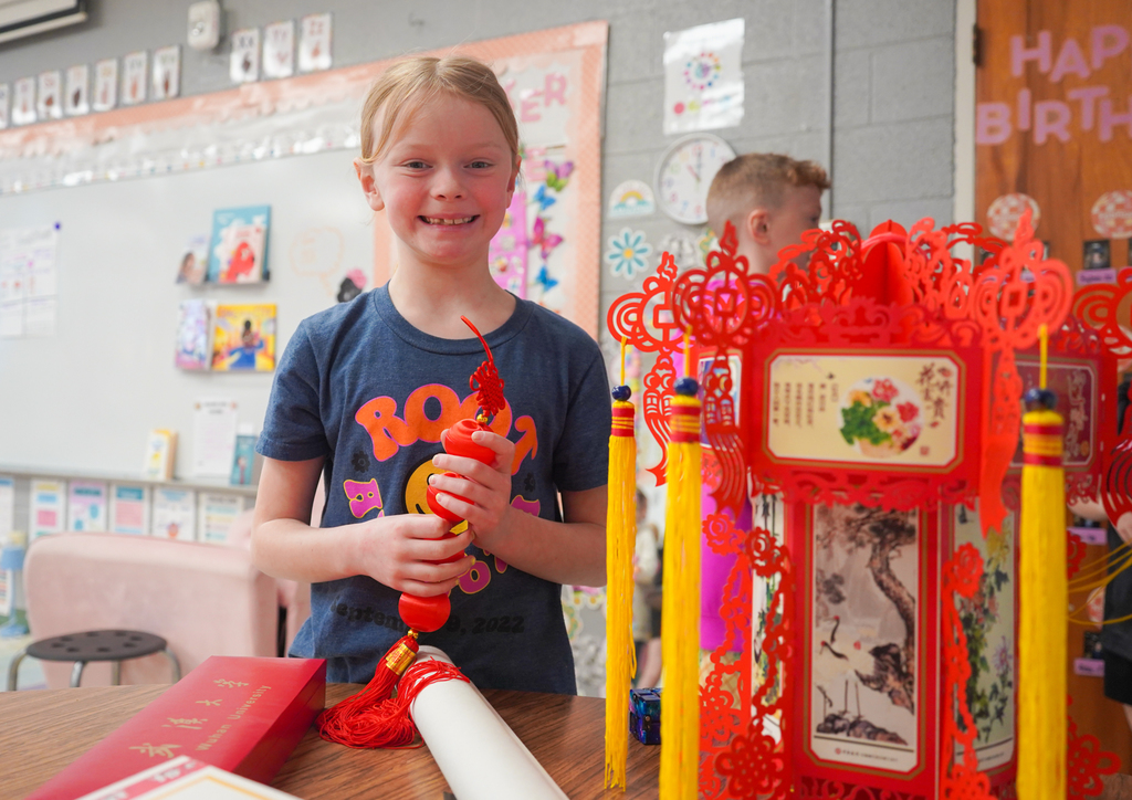 A girl smiling, holding a red lantern with tassels. She stands in front of a desk with a red lantern and paper crafts.
