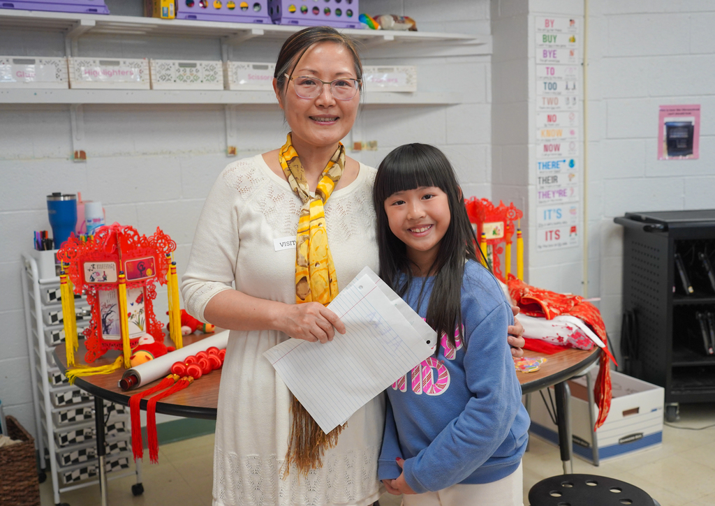 Woman with glasses and a yellow scarf stands next to a girl holding a paper, in a room.