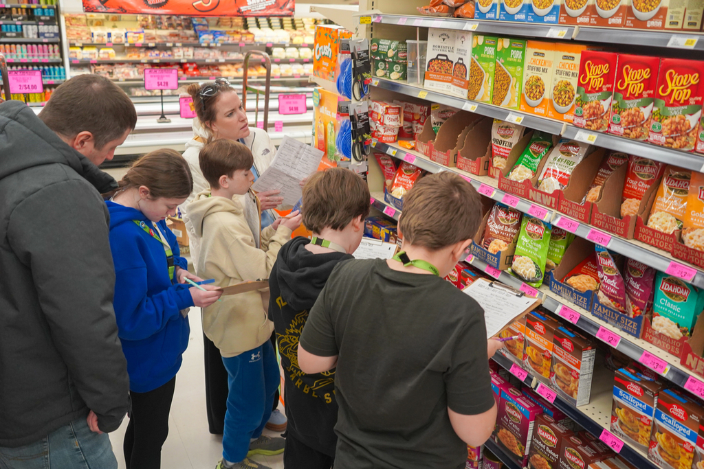 A family examines items on a grocery aisle, with several children holding clipboards. They appear to be reading product labels.