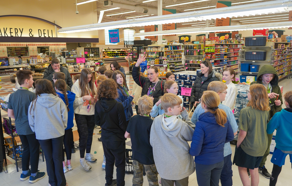 A group of students, likely in a school setting, gather in a supermarket. Some hold shopping carts.