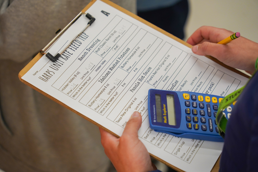 Person holds clipboard with checklist and blue calculator. Paper has text and numbers, including "Maths" and "English".