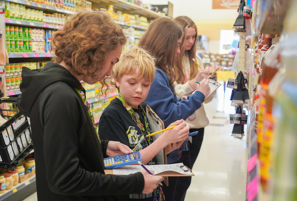 Several children in a store aisle are looking at items on the shelves and taking notes.