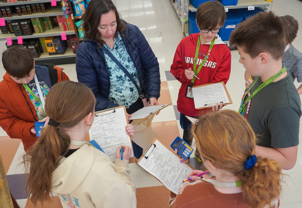 Children with clipboards in a school store. A teacher stands with them. Background shows shelves of products.
