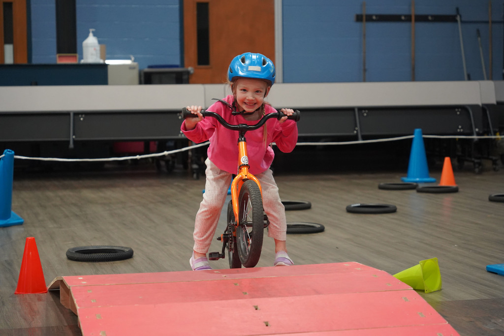 Child in pink top and helmet riding small bike with training cones on wooden floor.
