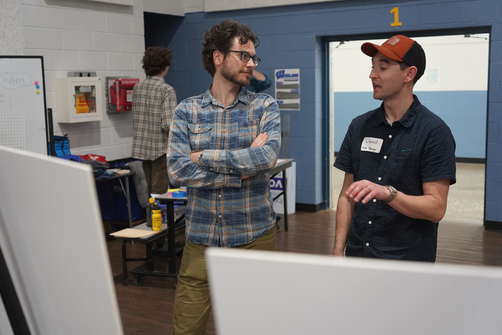 Two men in a room with blue walls. One wears a plaid shirt; the other, a cap. They converse.
