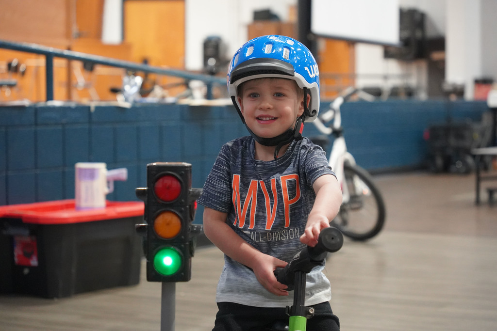 A young child rides a scooter with a green light signal. He wears a helmet. A storage box and a traffic light are behind him.