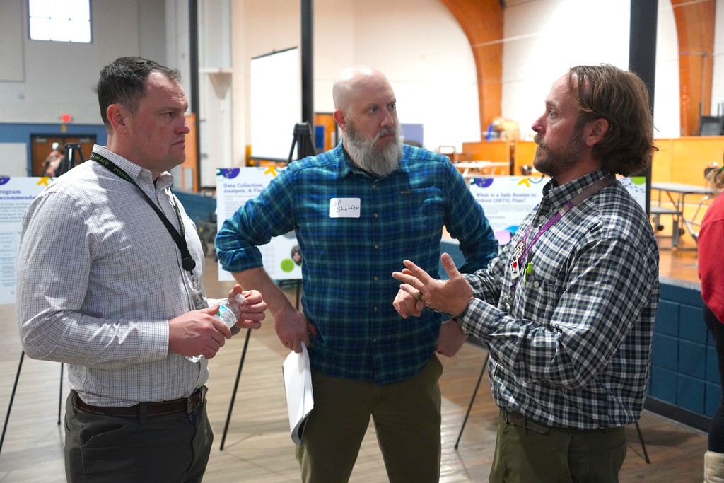 Three men converse in an indoor area with large windows, a stage, and tables in the background.