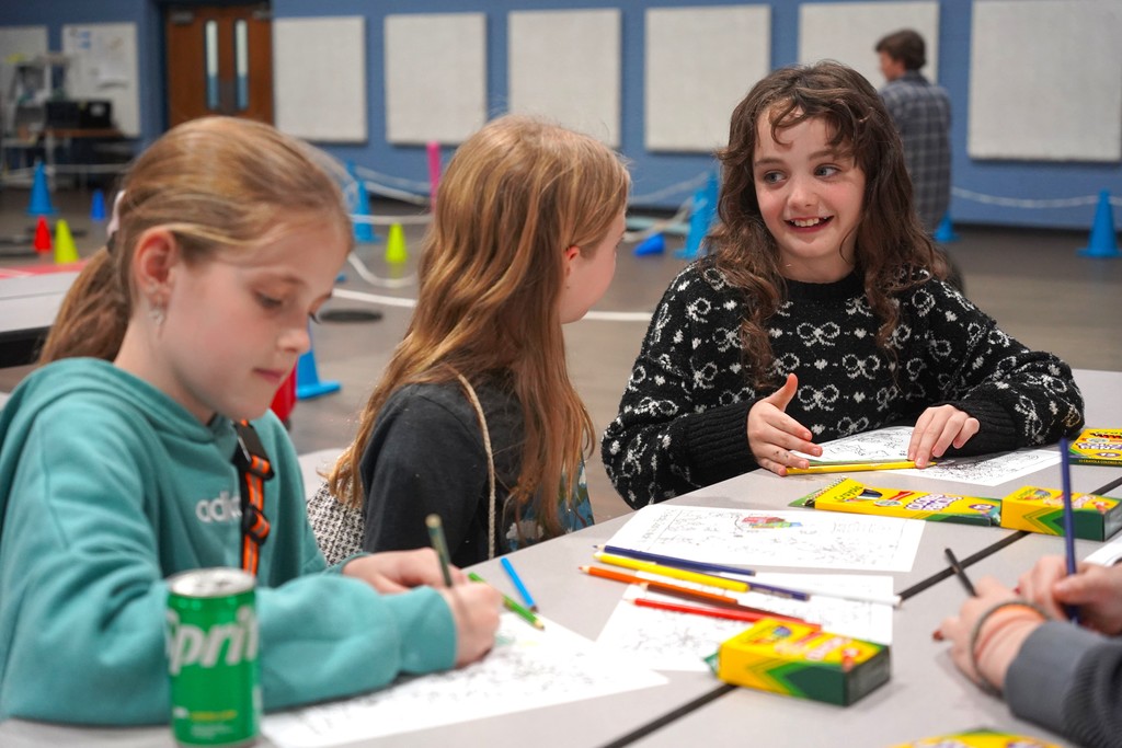 Three girls in a classroom, engaged in coloring with pencils and markers on paper. A green can is on the table.
