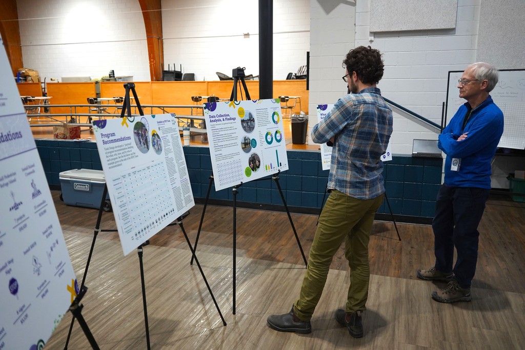 Two people view posters on a stand in a large room with wooden floors and a bleacher.