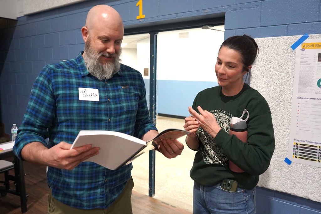 A man and woman examine a document while standing in a room with blue walls and a white board.