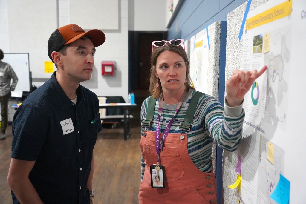 A man and a woman stand by a bulletin board with various papers. The woman points at a paper.
