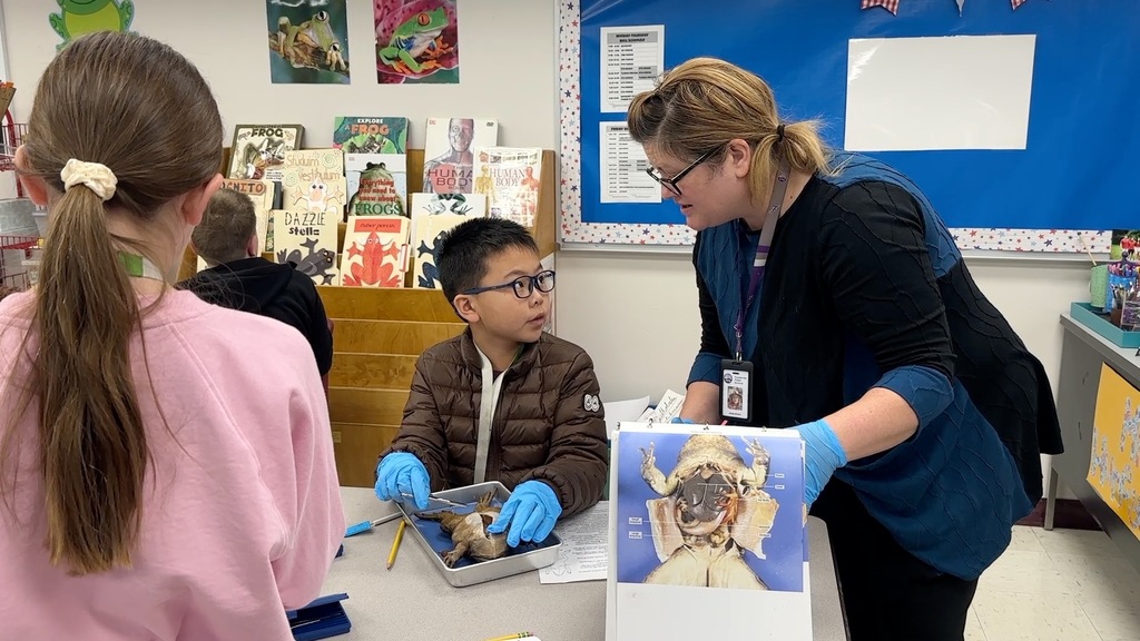 A woman in blue gloves examines an object while two children watch. A classroom with bookshelves and a blue bulletin board is in the background.