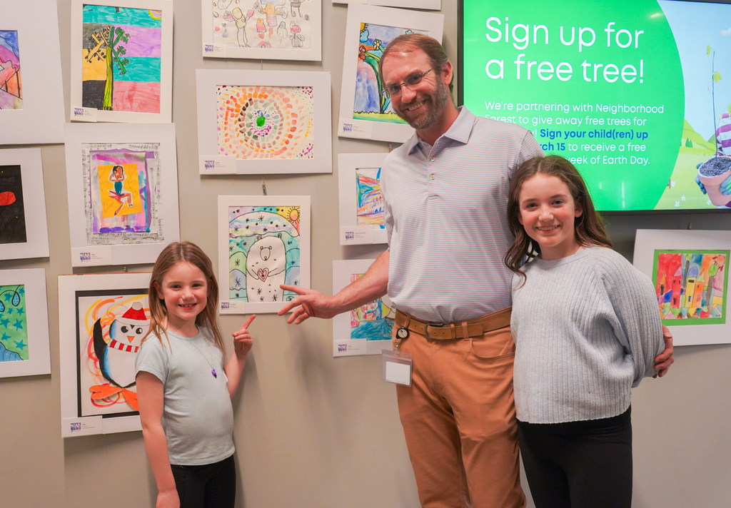 A man and two girls pose in front of a wall of children's artwork.