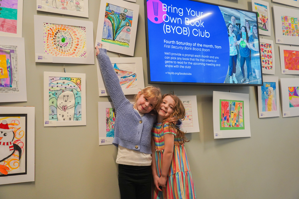 Two children posing for a photo in front of a wall with framed artwork and a large screen.